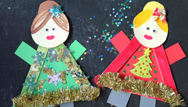 Two paper dolls dressed in festive holiday outfits, each featuring an ugly sweater craft made with colored paper, tinsel, sequins, and glitter. One wears green with snowflakes, the other red with a Christmas tree—both smiling and accessorized.