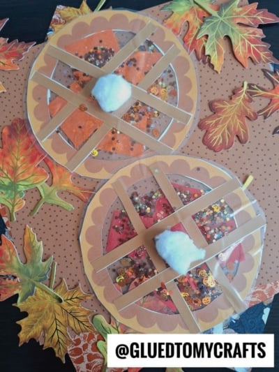 Two round, paper craft stained glass pies decorated with tan lattice strips, brown scalloped edges, clear sequin-filled sections, and cotton whipped cream centers. Autumn leaf decorations surround them. @GLUEDTOMYCRAFTS is written at the bottom.
