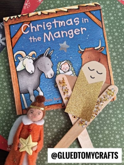 A Christmas book titled Christmas in the Manger is displayed with a handmade craft stick baby Jesus, an angel craft made of craft sticks, paper, and glitter, plus a felt angel doll. The handle @GLUEDTOMYCRAFTS is shown at the bottom.