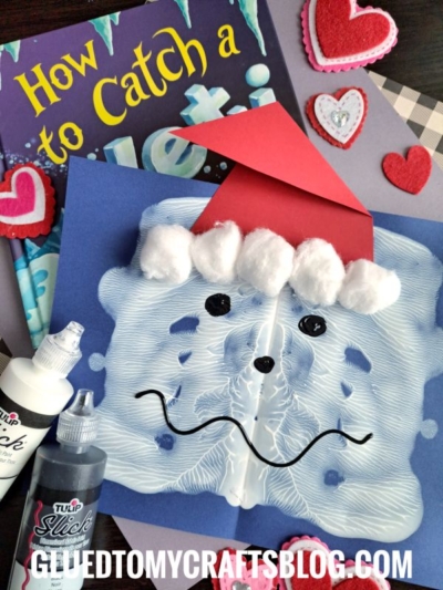 A childrenās craft project features a blue paper with white paint smeared to resemble Santaās faceālike a festive paint splat yetiāwith cotton balls for his hat trim, a red paper hat, and a black drawn smile, surrounded by books and heart decorations.