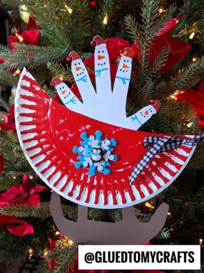 A festive paper plate snowman sleigh craft, featuring a red painted sleigh with a handprint of snowmen as passengers, decorated with a snowflake and ribbon, and perfect for displaying on your Christmas tree.