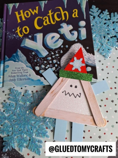 A children’s book titled How to Catch a Yeti sits beside a popsicle stick Christmas yeti craft made from paper and glitter, arranged on a table with blue snowflake decorations. @GLUEDTOMYCRAFTS is written at the bottom.