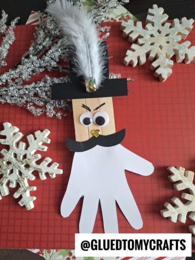 A popsicle stick nutcracker-inspired snowman craft featuring googly eyes, a black paper hat and mustache, gold bead nose, white feather, and handprint body. Finished with snowflakes on a red plaid background. @GLUEDTOMYCRAFTS at the bottom.