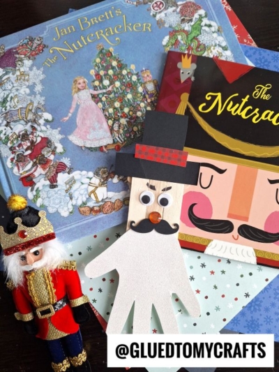Children’s Nutcracker-themed books, a Nutcracker figurine, a popsicle stick nutcracker, and a handmade Nutcracker craft made from a traced handprint with googly eyes are arranged on a table. The handle @GLUEDTOMYCRAFTS is displayed.