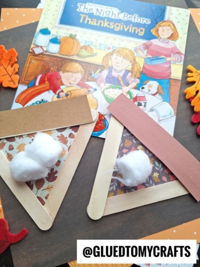 Two Thanksgiving-themed crafts, including a popsicle stick apple pie made with brown and red paper and cotton balls, sit on top of a children’s book titled The Night Before Thanksgiving. Felt leaves decorate the scene. Text reads @GLUEDTOMYCRAFTS.