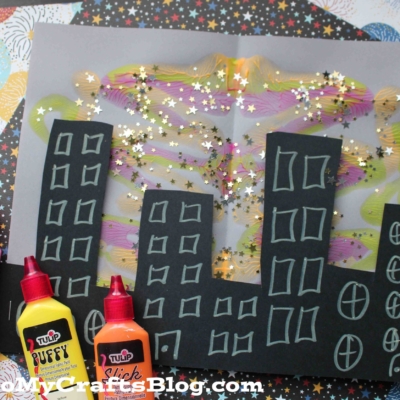 A colorful children’s craft features a new year's eve city skyline on black paper with yellow windows, painted fireworks in the background, star-shaped glitter, and three bottles of Tulip paint.