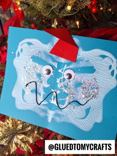 A blue paper craft of a silly paint splat yeti face with googly eyes, white paint, glitter, a red paper hat, and a squiggly black mouth is displayed on a Christmas tree. @GLUEDTOMYCRAFTS is written at the bottom.