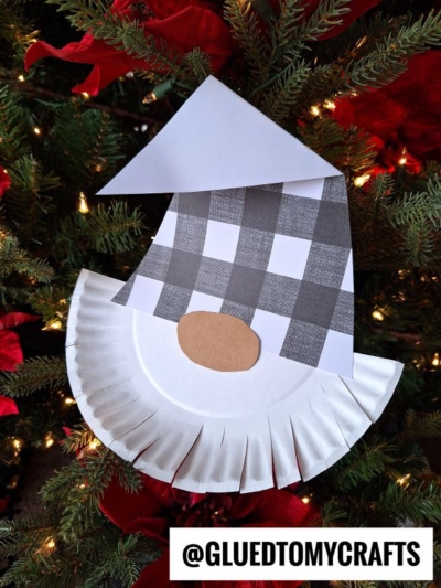 A handmade paper plate bearded Christmas gnome ornament, featuring a patterned paper hat and oval nose, hangs on a festive tree adorned with red poinsettias and lights. @GLUEDTOMYCRAFTS is displayed at the bottom.
