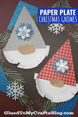 Two paper plate bearded Christmas gnome crafts with white beards, oval noses, and patterned hats (one gray with dots, one red plaid), surrounded by snowflake decorations on a dark background.