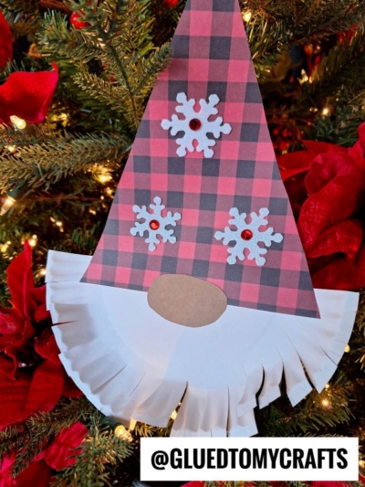 A handmade paper plate bearded Christmas gnome ornament with a plaid hat, paper snowflakes, and a fringed white beard hangs on a tree decorated with poinsettias and twinkling lights. @GLUEDTOMYCRAFTS appears at the bottom.