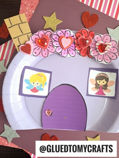 A handmade paper plate valentine fairy house craft made from a purple plate, decorated with paper hearts, flowers, and glitter stars. Two cartoon fairies peek from the windows, and a heart adorns the purple door.