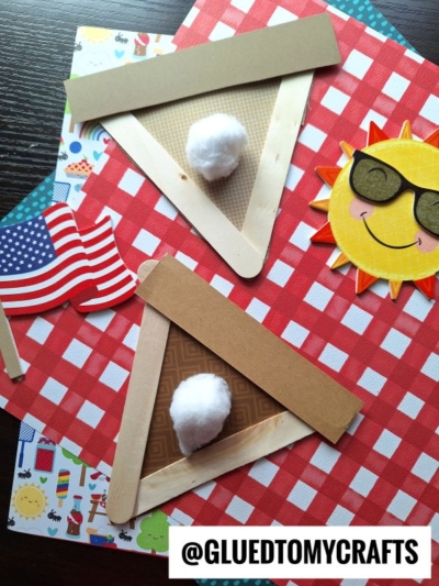 Two popsicle stick apple pie crafts with cotton ball "whipped cream" sit on red checkered paper. Nearby are a smiling sun with sunglasses, an American flag cutout, and colorful patterned paper. Text reads @GLUEDTOMYCRAFTS.