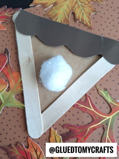 A craft stick apple pie slice made from craft sticks, brown paper, and a cotton ball, displayed on a fall-themed, leaf-patterned background.