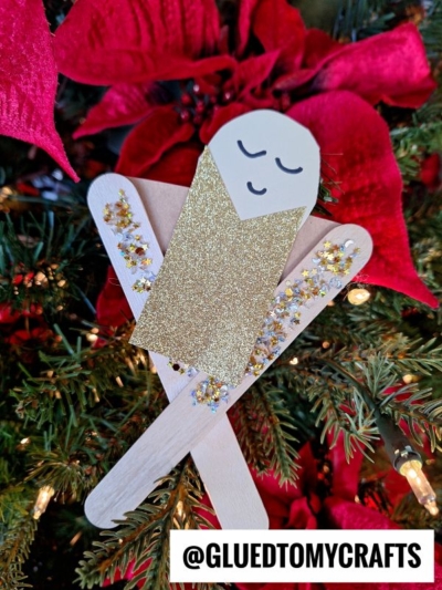 A Popsicle Stick Baby Jesus In A Manger ornament, made from craft sticks, gold glitter paper, and cardstock, hangs on a Christmas tree with red poinsettia flowers and twinkling lights. Text at bottom reads “@GLUEDTOMYCRAFTS.”.