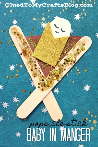 A Popsicle Stick Baby Jesus In A Manger craft shows a gold paper baby wrapped in a blanket, lying in a manger made of overlapping popsicle sticks and glitter on a brown paper background with star confetti.