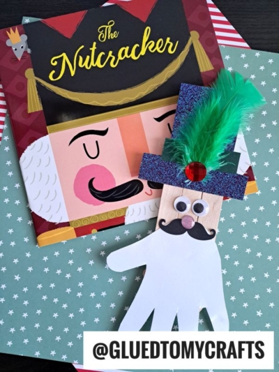 A Nutcracker book rests on star-patterned paper beside a handprint craft and a popsicle stick nutcracker, both decorated with googly eyes, a mustache, glittery hat, green feather, and red gem. @GLUEDTOMYCRAFTS is at the bottom.