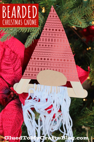 A handmade popsicle stick bearded Christmas Gnome ornament with a red patterned hat and white yarn beard, hanging on a Christmas tree adorned with red poinsettias.
