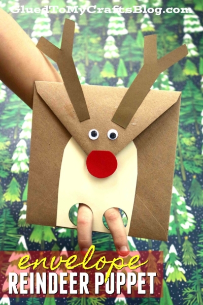 A hand holds a reindeer envelope puppet with googly eyes, brown antlers, a red nose, and finger holes for legs, set against a festive forest background. Text reads reindeer envelope puppet.