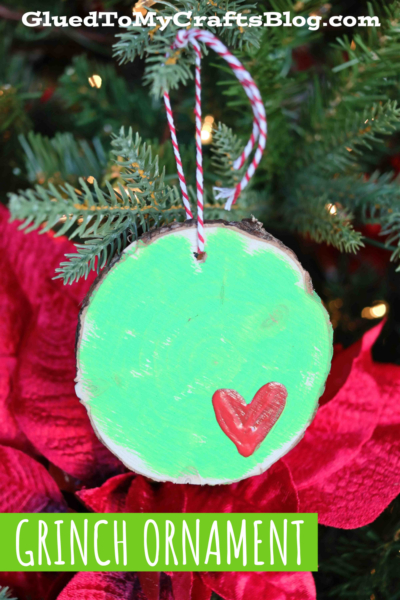 A round wood slice Grinch ornament painted green with a red heart hangs on a Christmas tree with red and white string. Red poinsettias and pine branches fill the background. Text reads “GluedToMyCraftsBlog.com” and “GRINCH ORNAMENT.”.