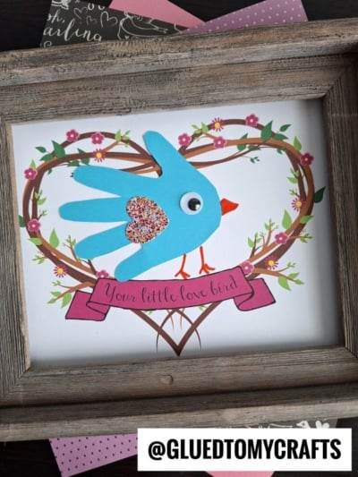 A framed craft featuring a blue handprint "my little love bird" cutout with a googly eye and glitter, perched on a heart-shaped branch with pink flowers. A pink banner reads Your little love bird. @GLUEDTOMYCRAFTS is at the bottom.