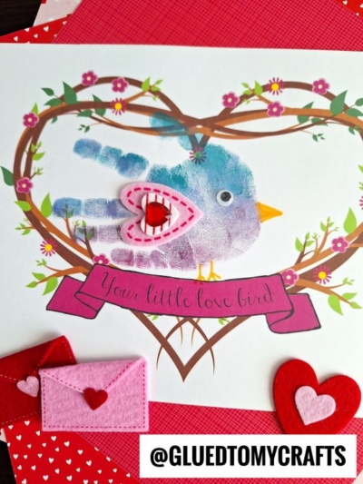 A craft featuring a blue and purple handprint my little love bird with a pink felt heart wing inside a floral heart wreath, topped with a banner reading Your little love bird. Felt hearts and mini envelopes accent the bottom.