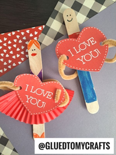 Two Valentine Puppet Friends made from craft sticks hold red heart signs that say “I LOVE YOU!” One wears a red cupcake liner skirt and dress; the other has blue pants. Both have pipe cleaner arms. @GLUEDTOMYCRAFTS is shown below.