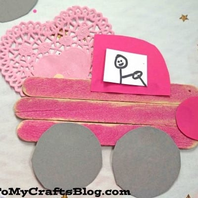 A popsicle stick valentine love truck craft made from pink popsicle sticks, heart-shaped paper doilies, gray paper wheels, and a pink cab with a hand-drawn figure, displayed on a white surface with gold stars.