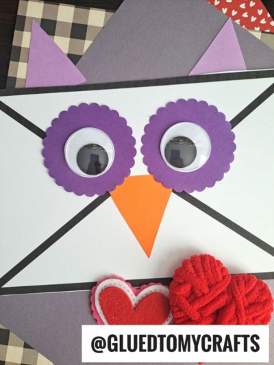A handmade Envelope Owl Card crafted from an envelope with large googly eyes, a purple scalloped circle around each eye, a triangle orange beak, and heart decorations, labeled @GLUEDTOMYCRAFTS at the bottom.