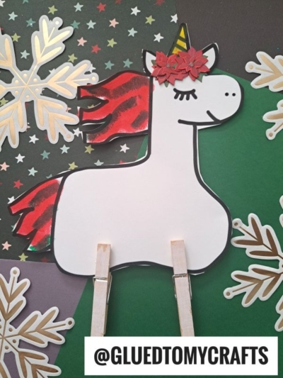 A whimsical clothespin unicorn craft featuring a red glitter mane, a flower crown, and clothespin legs is displayed on a festive background adorned with gold snowflakes. @GLUEDTOMYCRAFTS is written at the bottom.