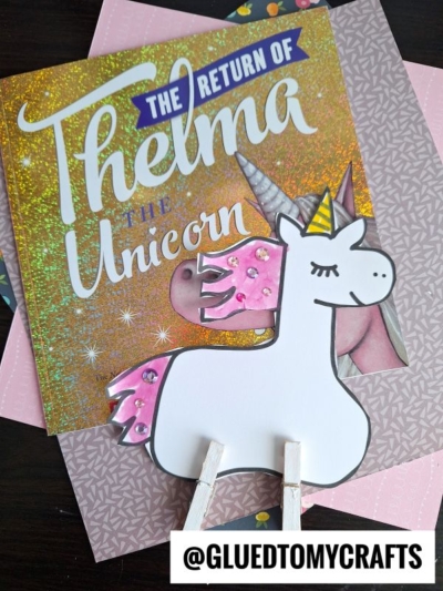 A charming clothespin unicorn craft with pink details stands atop the glittery book The Return of Thelma the Unicorn. A watermark reads @GLUEDTOMYCRAFTS.