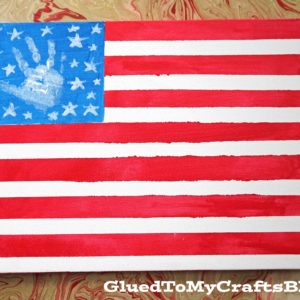 A handmade handprint flag painting features a blue rectangle with white stars and a white handprint, plus red and white stripes. The marbled background adds flair. The bottom right reads GluedToMyCraftsBlog.com.