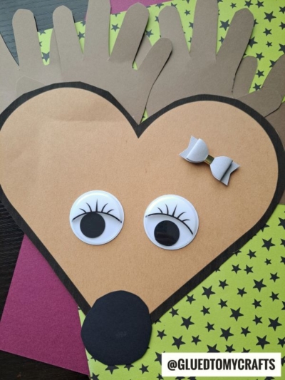 A reindeer craft made from brown paper shaped like a heart, with large googly eyes, a black nose, antlers, and a small white bow on its head—much like the charming paper handprint hedgehog projects kids love—all set against a background with star patterns. @GLUEDTOMYCRAFTS.