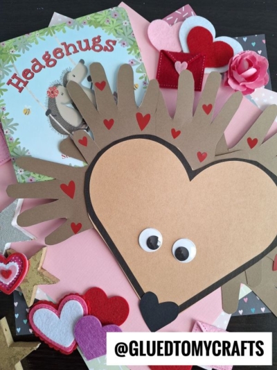 A handmade paper handprint hedgehog craft made from heart-shaped brown paper, googly eyes, and red heart stickers is surrounded by felt hearts and the children's book Hedgehugs. @GLUEDTOMYCRAFTS is written at the bottom.
