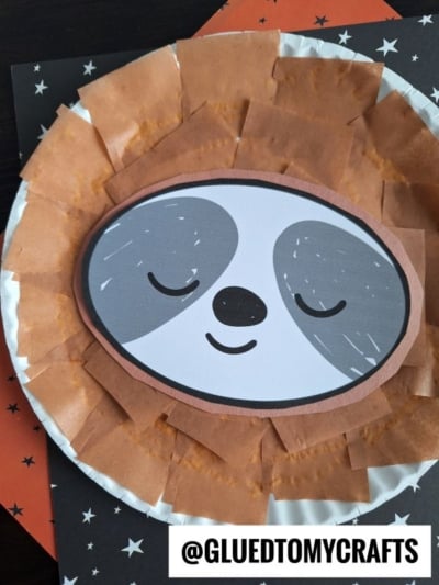 This paper plate sloth craft features a cartoon sloth face in the center, brown tissue paper edging, and a starry black background with an orange accent—perfect for a fun and easy paper plate sloth activity!.