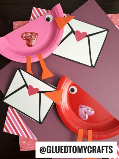 Two paper plate valentine love note birds, one pink and one red, hold envelope-shaped cards with heart seals. The scene is set on patterned and solid colored paper, with a @GLUEDTOMYCRAFTS watermark at the bottom.