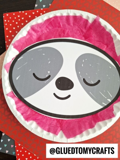 A paper plate sloth craft featuring a smiling sloth face with closed eyes, gray patches, and pink tissue paper around the edge. The background includes heart-patterned papers and the text @GLUEDTOMYCRAFTS.