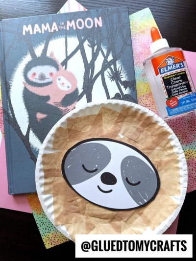 A children’s book titled Mama in the Moon, a bottle of Elmer’s glue, and a paper plate valentine sloth craft made from colored paper are arranged on colorful sheets. The handle @GLUEDTOMYCRAFTS is at the bottom.