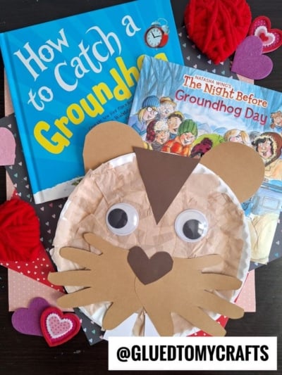 Two children’s books about Groundhog Day, heart-shaped felt decorations, and a cute paper plate groundhog craft with googly eyes are arranged on a table. @GLUETOMYCRAFTS appears at the bottom.