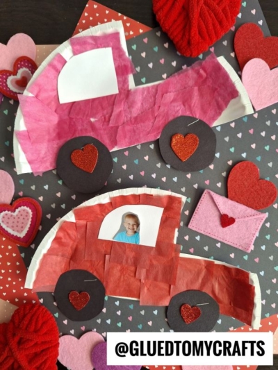 Two love bug cars crafted from tissue paper, hearts, and a child's photo are displayed on a black background decorated with red, pink, and white hearts. An envelope and heart embellishments surround each paper plate love bug creation.