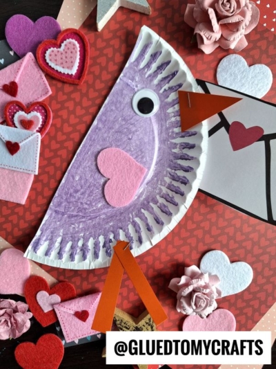 A purple paper plate is crafted into a valentine love note bird with a beak, eye, wings, and heart details, surrounded by felt hearts, paper flowers, and tiny envelopes on a red heart-patterned background. @GLUEDTOMYCRAFTS is labeled below.