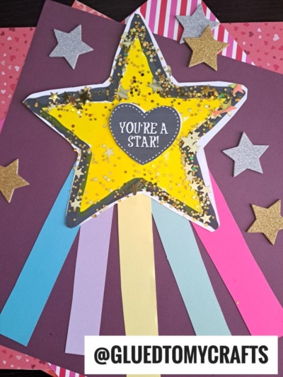 A handmade You're A Star Paper Badge, star-shaped with "You're a Star!" in the center, decorated with gold glitter, surrounded by glittery stars and colorful paper strips. The handle @GLUEDTOMYCRAFTS is at the bottom.