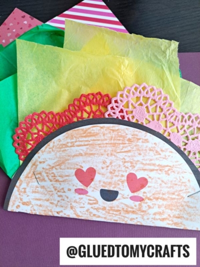 A handmade paper love taco craft with a smiling face and heart eyes, featuring green and yellow tissue paper for lettuce and cheese, plus pink paper doilies as toppings. The handle @GLUEDTOMYCRAFTS is at the bottom.