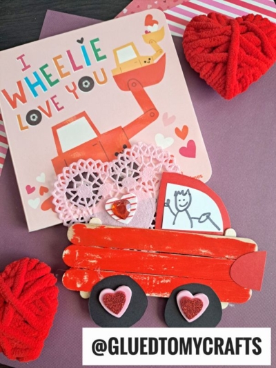 A cute popsicle stick valentine card with a red love truck and “I Wheelie Love You,” surrounded by red heart yarn decorations. @GluedToMyCrafts is written at the bottom.