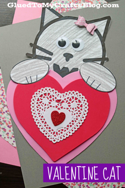 A Valentine heart cat craft featuring a gray cat made of cut-out paper shapes, holding a large red and pink heart adorned with a white doily and a glittery red heart. The valentine cat is finished with a cute pink bow by its ear.