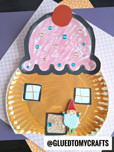 A paper plate cupcake house craft shaped like a cupcake, featuring a painted plate, hand-drawn door, windows, pink frosting, sequins, and a red cherry. A small gnome figure stands by the door. @GLUEDTOMYCRAFTS is written at the bottom.