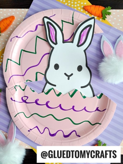 A paper plate craft shows a peeking Easter bunny emerging from a decorated pink egg with zigzag patterns. Fake carrots and bunny ears with fluffy tails are arranged on a striped surface, capturing the fun of a peek a boo Easter bunny.