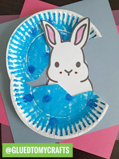 A paper plate painted blue is cut and shaped like an Easter egg, opening to reveal a cute, hand-drawn peek a boo easter bunny with pink ears and cheeks. The background includes pink and gray papers. @GLUEDTOMYCRAFTS is written in green.