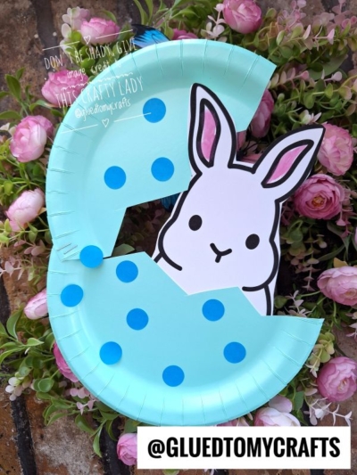 A light blue paper plate decorated with blue dots is cut to resemble the letter C and features a peeking Easter bunny illustration in the center, surrounded by pink flowers. The handle @GLUEDTOMYCRAFTS appears at the bottom.