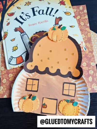 A fall-themed children’s book titled Its Fall! sits behind a paper plate cupcake house craft shaped like a brown scoop, decorated with paper pumpkins, gold dots, and small windows. @GLUEDTOMYCRAFTS is watermarked on the image.