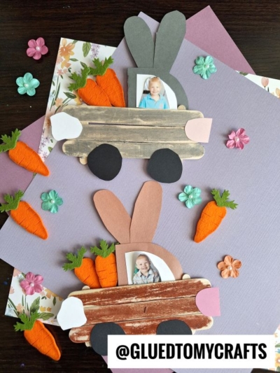 Two handmade craft stick bunny truck crafts display children's photos, decorated with orange carrots, flowers, and pastel backgrounds. Made from paper and popsicle sticks, each features a unique touch. The @GLUEDTOMYCRAFTS watermark is at the bottom.